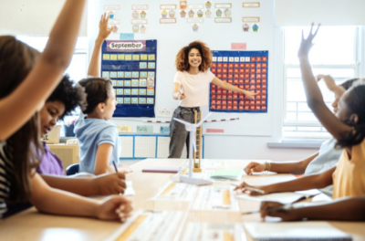 A smiling teacher calls on students who are raising their hands.